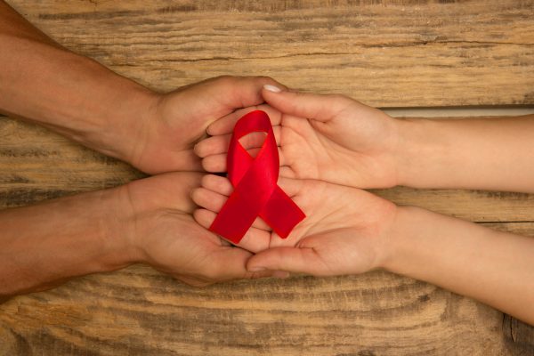 Female and male hands holding red HIV and AIDS awareness ribbon isolated on wooden background. Concept of healthcare and medicine, worldwide supporting, volunteer campaign against illness.